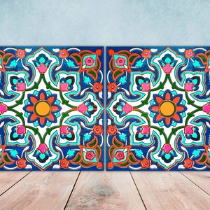 May include: Two square ceramic tiles with a symmetrical, intricate design featuring a central flower with orange, red, and yellow petals surrounded by blue, green, and pink floral motifs. The tiles have a white background and a dark blue border.