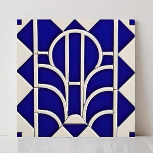 May include: Square ceramic tile with an Art Deco design. The tile has a vibrant blue background with a white, stylized floral or fan-shaped pattern. The design is symmetrical and the tile is surrounded by white and blue geometric shapes.