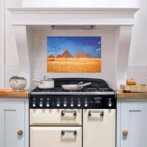 May include: A kitchen range with a black cooktop and cream-colored oven doors. Above the range is a tile backsplash with a framed print of the pyramids in Egypt. A colander with vegetables and a pot are on the countertop.