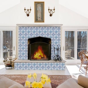 Puede incluir: Una habitaci&oacute;n blanca con una chimenea con azulejos de patrones azules y blancos. La chimenea tiene un marco de metal negro y un fuego ardiendo en su interior. Hay dos ventanas a cada lado de la chimenea y una mesa de madera con sillas en el fondo.