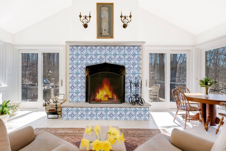 Puede incluir: Una habitaci&oacute;n blanca con una chimenea con azulejos de patrones azules y blancos. La chimenea tiene un marco de metal negro y un fuego ardiendo en su interior. Hay dos ventanas a cada lado de la chimenea y una mesa de madera con sillas en el fondo.
