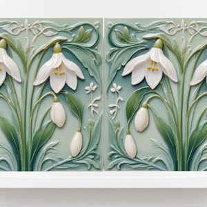 May include: Two square ceramic tiles with a floral design featuring white snowdrop flowers and green leaves against a pale green background. The tiles are displayed on a white shelf.