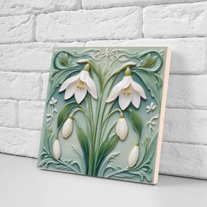 May include: A square, ceramic tile featuring a relief design of snowdrop flowers. The flowers are white with yellow centers, set against a pale green background with darker green leaves and stems. The tile has a light pink edge and is displayed against a white brick wall.