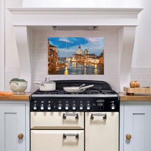 May include: A white kitchen with a black and cream range stove. The backsplash is decorated with a tile mural of a cityscape with a canal and boats. The mural is a nighttime scene with a blue sky and yellow lights.