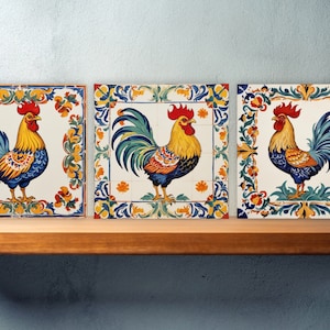May include: Three decorative ceramic tiles featuring a colorful rooster design. The roosters have yellow, blue, and orange plumage, with a red comb. Each tile is framed with a floral border in similar colors. The tiles are displayed on a wooden shelf.