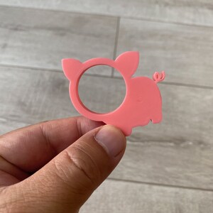 May include: Pink plastic pig napkin ring. The pig is in a standing position with its tail curled up. The ring is designed to hold a napkin in place.