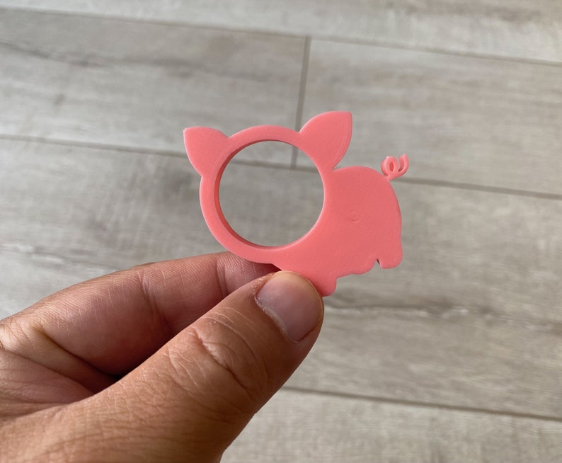 May include: Pink plastic pig napkin ring. The pig is in a standing position with its tail curled up. The ring is designed to hold a napkin in place.