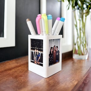 May include: A white, square pen holder with a photo on each side, holding several colorful markers. The pen holder sits on a wooden surface. The markers are white with pink, blue, green, and brown accents.