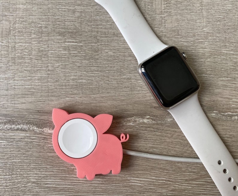 May include: A white Apple Watch with a black face and a pink pig-shaped charging stand.