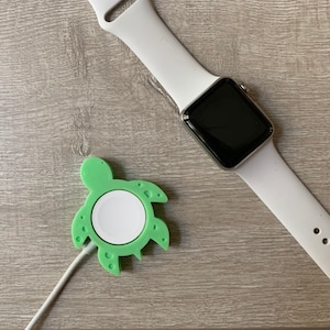 Può includere: Un supporto di ricarica a forma di tartaruga verde per un Apple Watch. Il supporto ha un pad di ricarica bianco e un cavo di ricarica bianco.