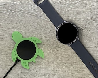 May include: A green turtle-shaped wireless charging pad with a black charging surface and a black charging cable. A black smartwatch with a black band is lying on a wood surface.
