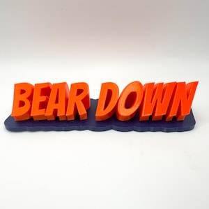 以下が含まれることがあります： 青いベースにオレンジ色の「BEAR DOWN」の文字。文字は太字で、ベースから際立っています。このアイテムはおそらく装飾品またはサインです。
