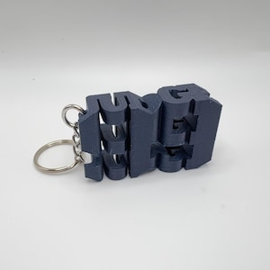 以下が含まれることがあります： 「JUNE」の文字が刻まれたネイビーブルーの3Dプリントキーホルダー。文字はつながっており、シルバーのキーリングに取り付けられています。キーホルダーは白い背景にあります。