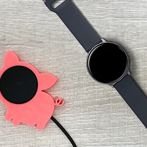 May include: A pink pig-shaped wireless charging pad with a black charging surface and a black charging cable. A black smartwatch with a black band is lying on a light wood surface.
