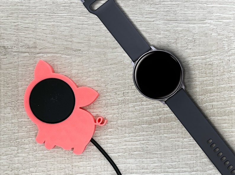 May include: A pink pig-shaped wireless charging pad with a black charging surface and a black charging cable. A black smartwatch with a black band is lying on a light wood surface.