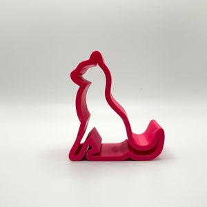 Puede incluir: Un soporte para teléfono de plástico rosa brillante con forma de perro sentado.