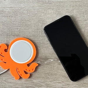 May include: An orange octopus-shaped wireless charging pad with a white charging surface. A black smartphone is lying on a wooden surface.