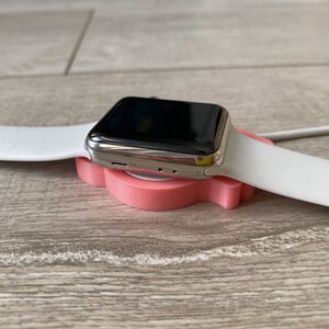 May include: A silver Apple Watch with a white band is resting on a pink charging stand.