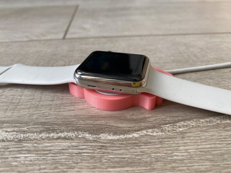 May include: A silver Apple Watch with a white band is resting on a pink charging stand.