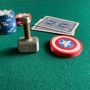 Op de afbeelding: Een zilveren en bruine speelgoedhamer met een triquetra-symbool op de kop, een rood-wit-blauw Captain America-schild en een stapel blauwe pokerchips op een groene vilt.