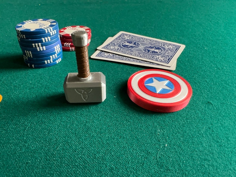 Op de afbeelding: Een zilveren en bruine speelgoedhamer met een triquetra-symbool op de kop, een rood-wit-blauw Captain America-schild en een stapel blauwe pokerchips op een groene vilt.