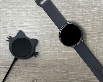 May include: A black smartwatch with a dark gray silicone band on a wooden surface. A black cat-shaped wireless charging stand is also on the surface.