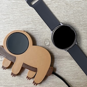 May include: A brown 3D printed sloth shaped charging stand with a black charging pad for a smartwatch. A black smartwatch with a black band is lying on a wooden surface.