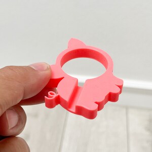 May include: A pink plastic pig-shaped phone holder. The holder has a hole in the middle for a phone to be placed.