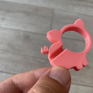 May include: A pink plastic pig-shaped ring holder. The ring holder has a hole in the middle for a ring to be placed.