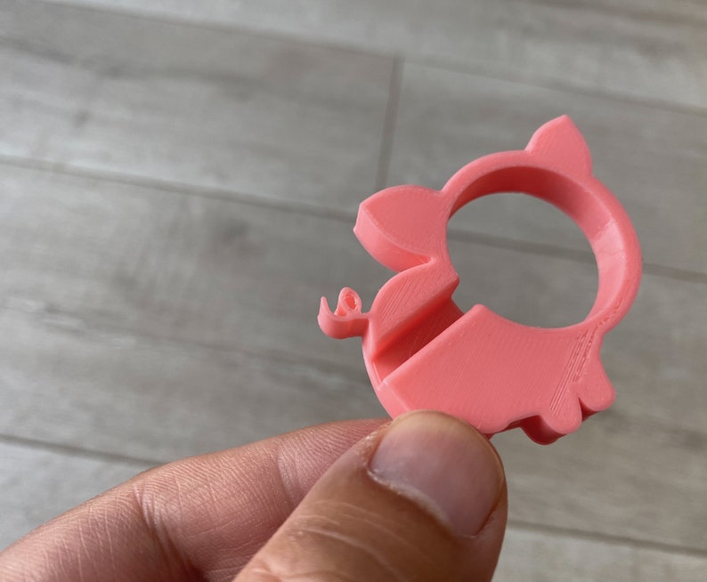 May include: A pink plastic pig-shaped ring holder. The ring holder has a hole in the middle for a ring to be placed.