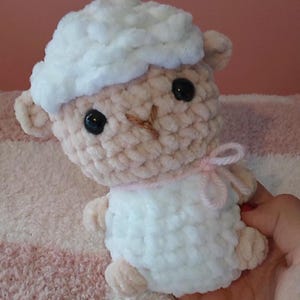 May include: A small, handmade crochet plush toy shaped like a lamb. The lamb features a fluffy white head and body, light beige limbs and face, two black plastic eyes, a stitched brown nose, and a thin light pink yarn bow tied around its neck.
