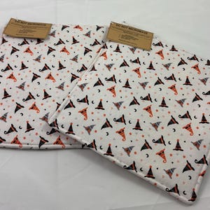 May include: Two square fabric hot pads featuring a white background with a repeating pattern of orange, black, and grey witch hats, stars, and crescent moons. Each pad has a brown rectangular tag attached that reads DO NOT MICROWAVE.