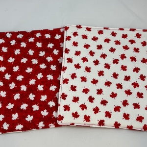 May include: Two square fabric pot holders featuring a maple leaf pattern. One pot holder has a red background with white maple leaves, while the other has a white background with red maple leaves. Both items have stitched edges and a flat design.