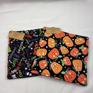 May include: Two square fabric bowl cozies featuring Halloween patterns. One displays orange jack-o'-lanterns on a black background with white stars, while the other shows colourful text including "Boo!" and "Fright" on black. Each has a brown tag.