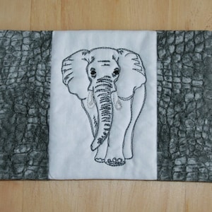May include: A white fabric coaster with a black embroidered elephant design. The coaster has a gray crocodile skin pattern on the sides.
