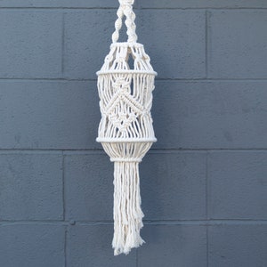 May include: A white macrame plant hanger with a wooden ring at the top. The hanger is made of knotted cord and has a long fringe at the bottom.