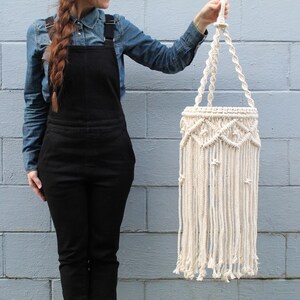 May include: A white macrame hanging planter with a geometric pattern and long fringe. The planter is being held by a person wearing a denim shirt and black overalls.