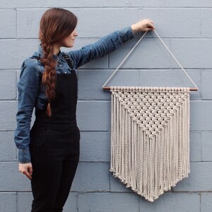 May include: A white macrame wall hanging with a triangular shape and fringe. The hanging is attached to a wooden dowel rod.