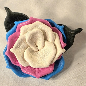 May include: A handcrafted leather flower in the colors of the bi pride flag. The flower features petals in blue, pink, white, and black. The petals are layered to create a realistic rose shape. The flower is a unique accessory.