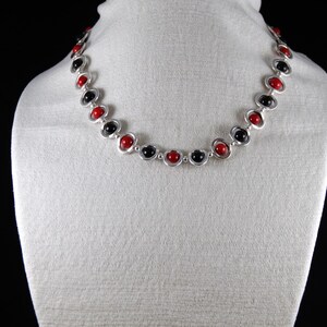 Puede incluir: Collar con cuentas rojas y negras alternadas, engarzadas en metal plateado. Las cuentas son redondas y están espaciadas uniformemente. El collar se exhibe sobre un maniquí blanco.
