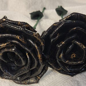 Puede incluir: Dos rosas de cuero negro con detalles dorados. Las rosas están dispuestas una al lado de la otra y tienen un aspecto realista.