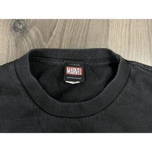 May include: Black t-shirt with a red Marvel logo tag attached to the neck. The tag says "Marvel" and "www.marvel.com".