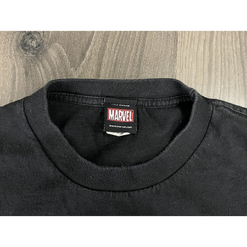 May include: Black t-shirt with a red Marvel logo tag attached to the neck. The tag says "Marvel" and "www.marvel.com".