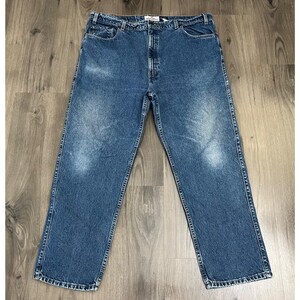 May include: Blue denim jeans with a classic five-pocket design. The jeans have a straight leg cut and a faded wash, with visible wear marks. The jeans are laid flat on a wooden surface, showcasing the front and back pockets and the belt loops.