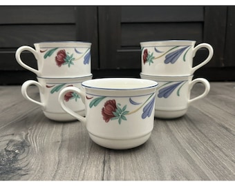 May include: A set of six white ceramic coffee mugs. Each mug features a floral design with red, blue, and green accents, and a blue trim. The mugs are stacked and arranged on a wooden surface.