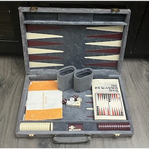 May include: Open backgammon set with a gray velvet interior, featuring ivory and burgundy playing fields. Includes dice, dice cups, game pieces, and rule books. The set is housed in a gray case with gold-tone hardware.