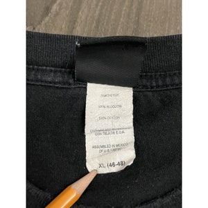 May include: Black t-shirt tag with white text. The tag says "RNI 0101531", "100% ALGODON", "100% COTTON", "ENSAMBLADO EN MEXICO CON TELA DE E.U.A.", "ASSEMBLED IN MEXICO OF U.S. FABRIC", and "XL (46-48)".