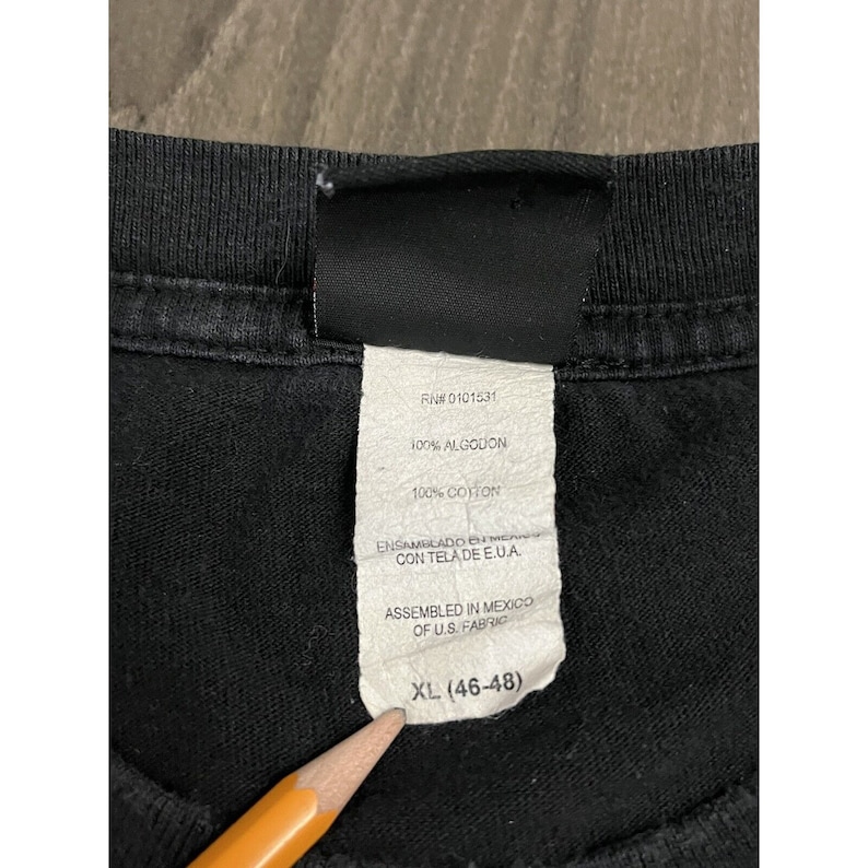May include: Black t-shirt tag with white text. The tag says "RNI 0101531", "100% ALGODON", "100% COTTON", "ENSAMBLADO EN MEXICO CON TELA DE E.U.A.", "ASSEMBLED IN MEXICO OF U.S. FABRIC", and "XL (46-48)".