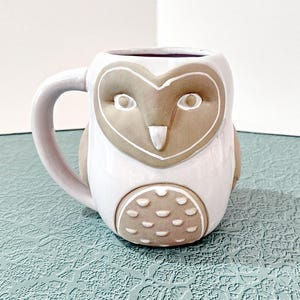 May include: A ceramic mug shaped like an owl with a white body and a light brown face and belly. The owl features a heart-shaped facial disc, a pointed beak, and small circular indentations on the belly. It has a curved handle on the left side.
