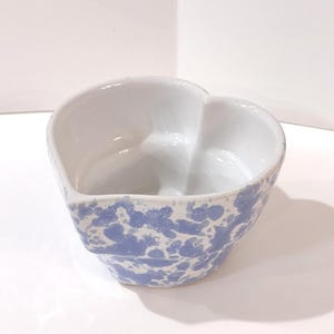 May include: A heart-shaped ceramic bowl with a white interior and a blue and white speckled exterior. The bowl is sitting on a white surface. The bowl is likely used for serving or decoration.